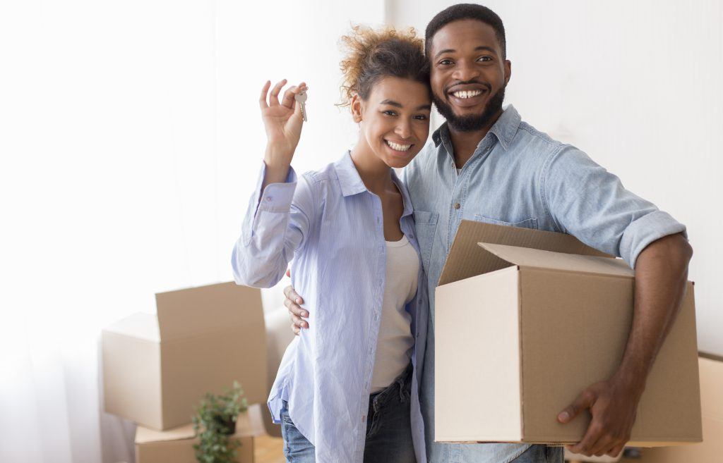 Own House. Happy Black Couple Showing Key Holding Moving Box And Hugging Standing In New Home.