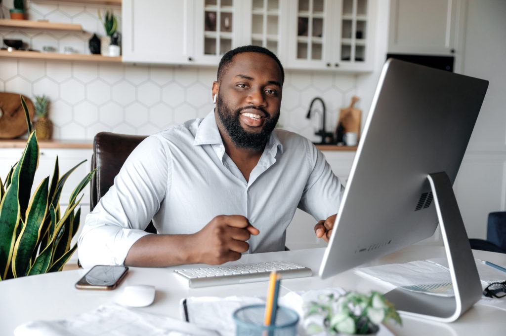 Portrait of a confident successful African American businessman, real estate agent, lawyer or manager, wearing a stylish shirt, working remotely at the computer, looking at camera, smiling friendly